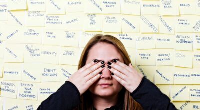 Kvinna med svarta naglar håller för sina ögon. Bakom henne mängder av postits med svåra ord.