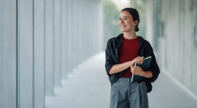 Kvinna med bok i handen står i korridor och blickar ut.