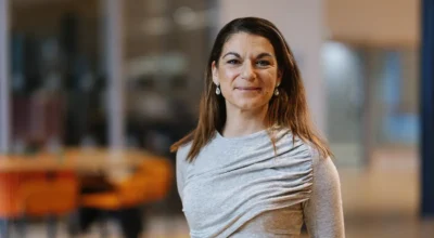 Kvinna i grå tröja tittar in i kameran och ler. Suddig bakgrund.