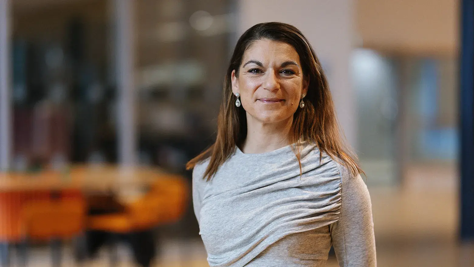 Kvinna i grå tröja tittar in i kameran och ler. Suddig bakgrund.